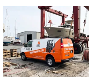 Orange and white mobile service vehicle at marina with red gantry cranes and sailboats in background