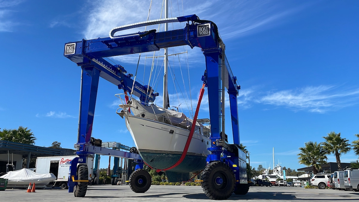 Blue gantry crane with red spreader bar hoisting white sailing yacht above concrete dock and marina basin