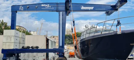 Blue gantry crane with yellow spreader bar hoisting dark-hulled powerboat over concrete dock and moored sailboats