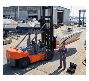 Technician in work attire with orange Toyota forklift in marine boatyard with sailboats and industrial sheds