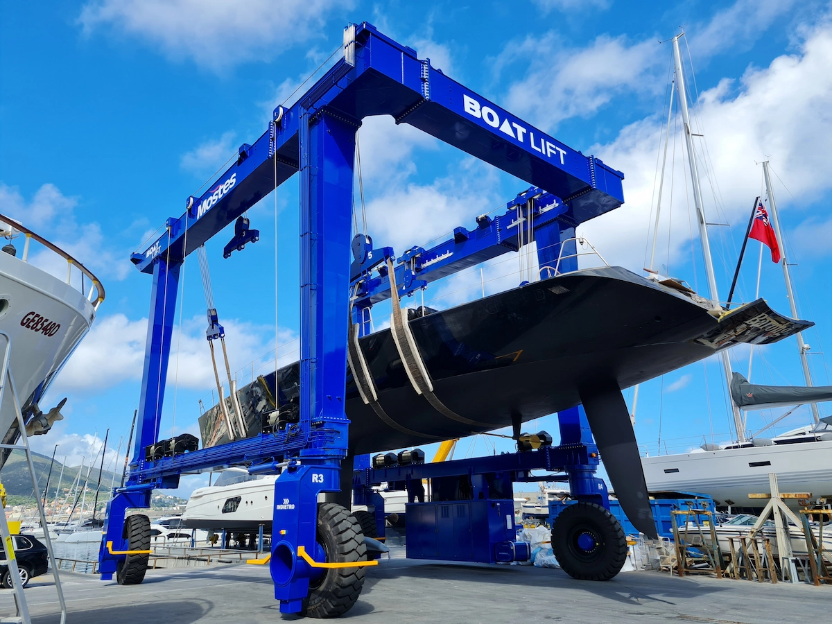 Blue gantry crane hoisting black-hulled sailing yacht with white cabin and red flag above marina basin with moored vessels