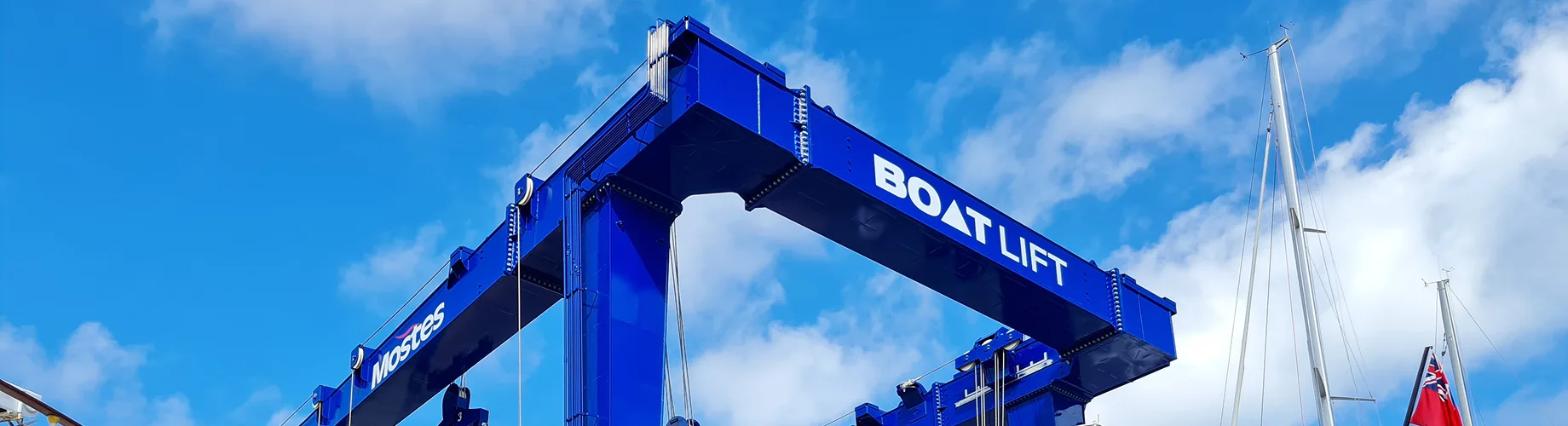 Blue gantry crane with "BOAT LIFT" text spanning twin columns against sky and sailing yacht masts