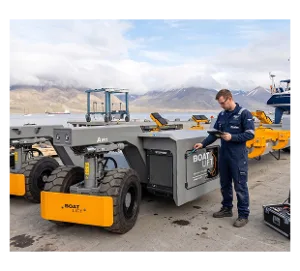 Technician in blue uniform servicing yellow and gray boat support cradle with mountain range and marina visible