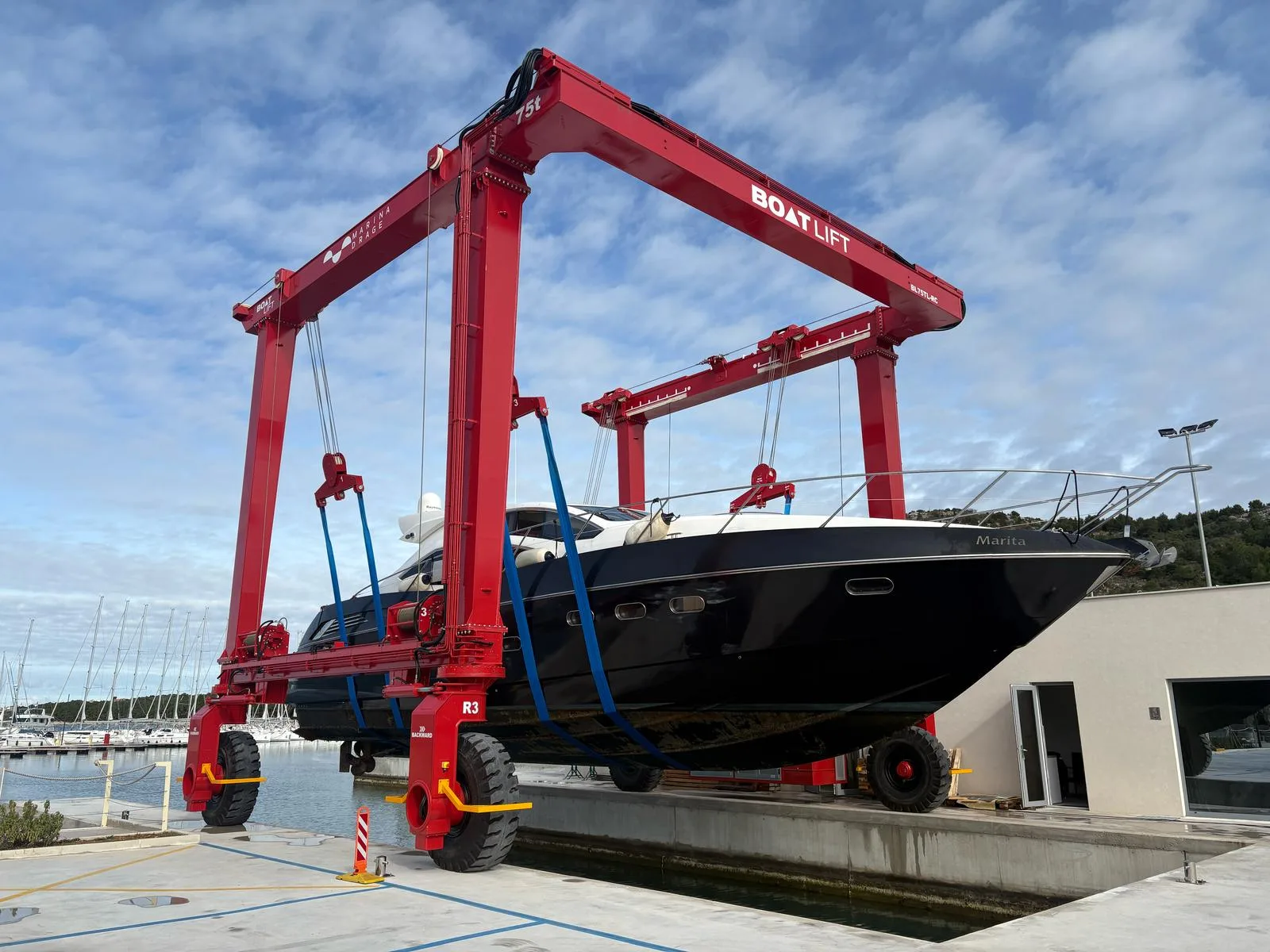 Red travel lift with blue straps hoisting black-hulled powerboat named Marita over concrete dock and marina basin